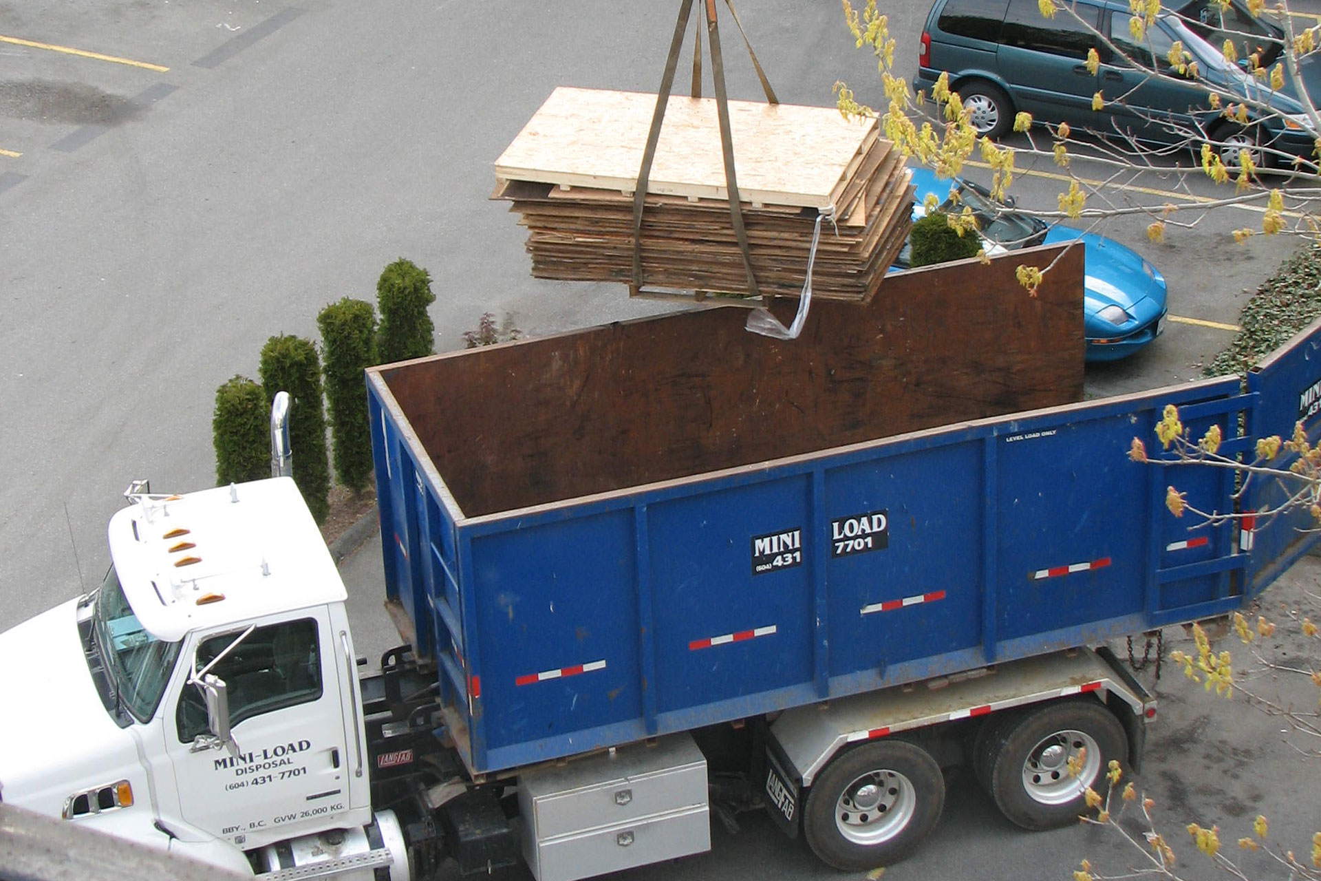 A crane lowers a stack of wooden boards into a blue dump truck parked in a lot next to a blue car and some small trees.