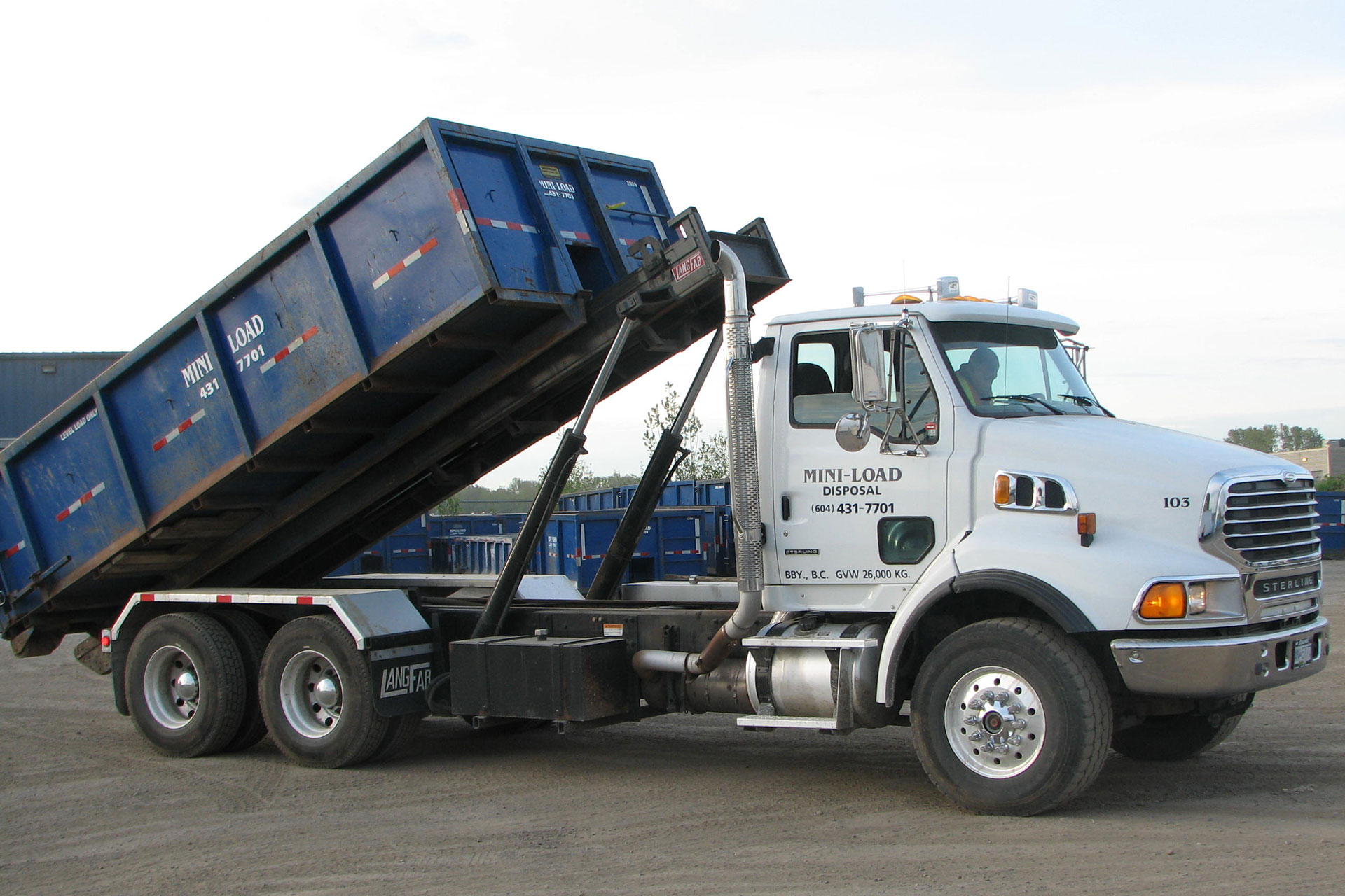 A white dump truck is lifting a large blue disposal bin at a work site.